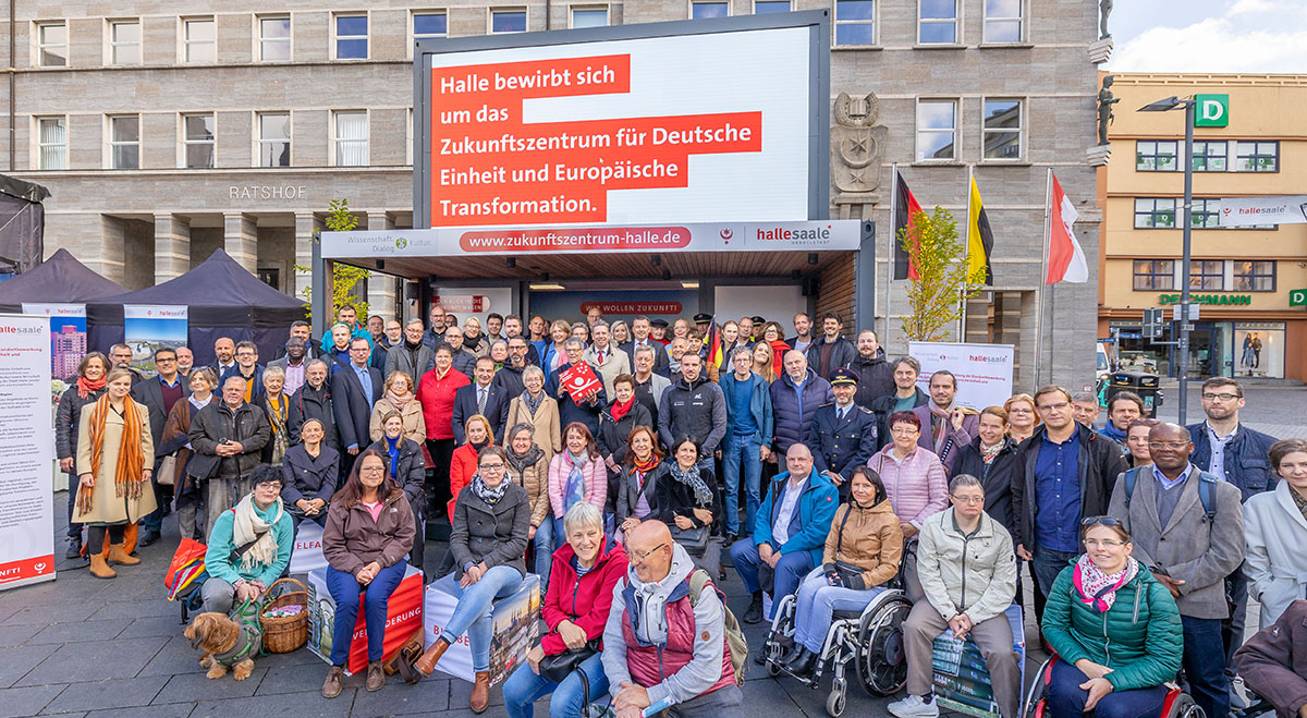 Viel Frauen und Münner - Gründungsmitglieder des Netzwerkes für das Zukunftszentrum in Halle auf dem Marktplatz vor einer Bühne vor dem Ratshof mit der Aufschrift Halle bewirbt sich um das Zukunftszentrum für Deutsche Einheit und Europäische Transformation
