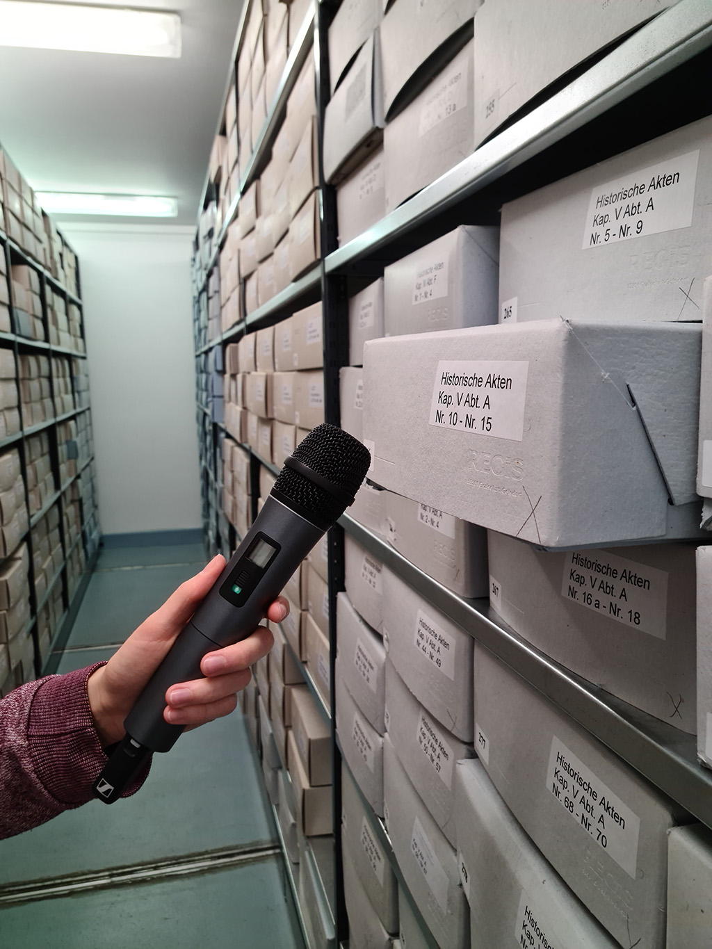 eine Hand mit Mikrofon vor einem aus dem Regal heraus ragenden Archivkarton mit historischen Akten