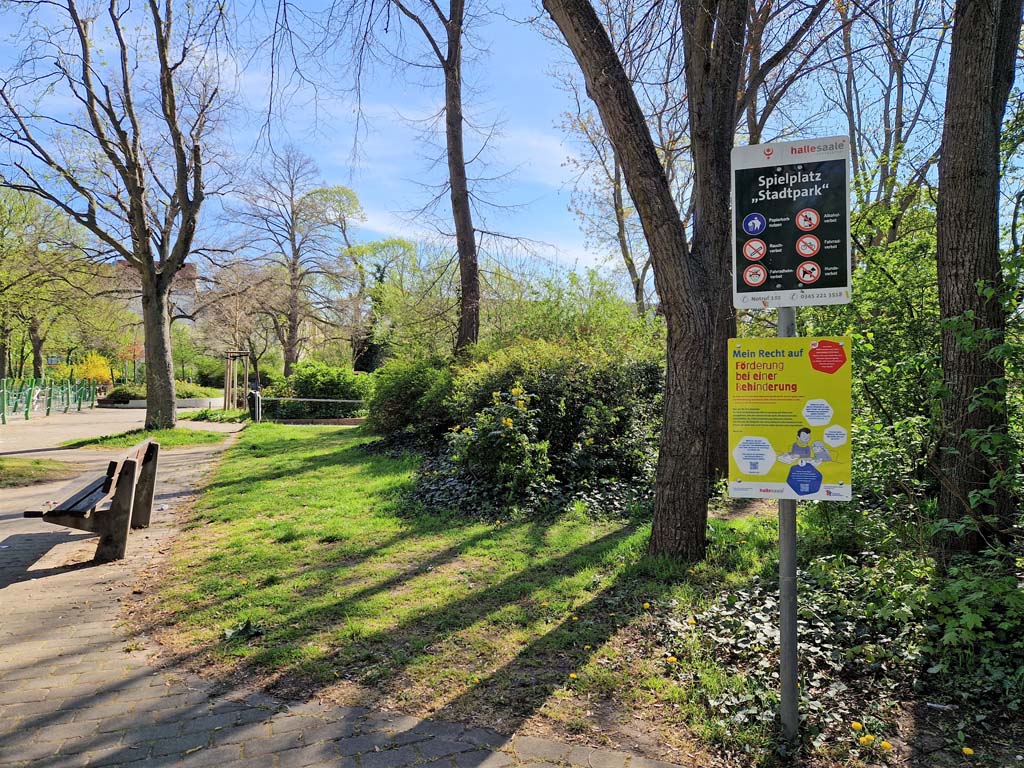 Station 9 - Spielplatz Stadtpark, Tafel zum Thema: Recht auf Förderung bei einer Behinderung am Wegrand dahinter sind Bäume