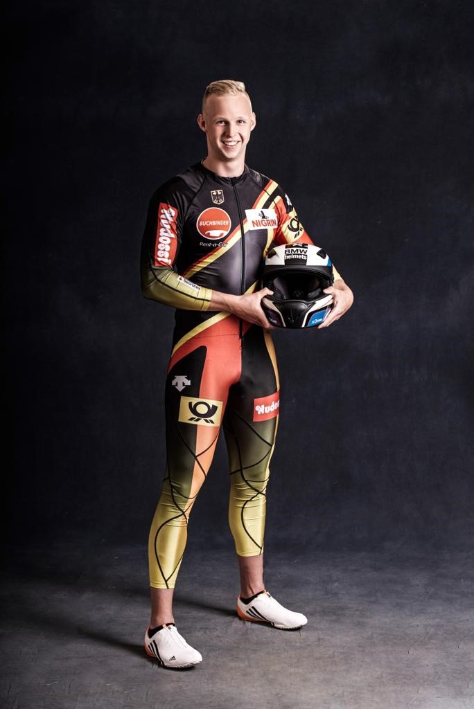 Der Sportler im Rennanzug. Seinen Helm hält er an der linken Seite an der Hüfte fest.
