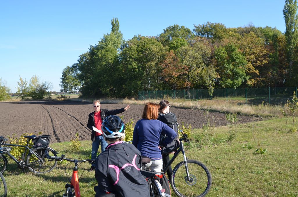 im Vordergrund steht eine Gruppe Radfahrer, im Hintergrund sieht man Bäume und Sträucher, ein Mann erläutert die Gehölzgruppe