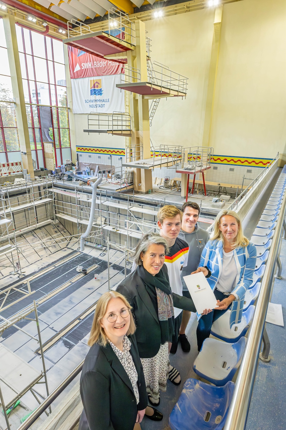 Die Personen stehen nebeneinander auf der Tribüne. Im Hintergrund das leere Becken mit Baugerüst.