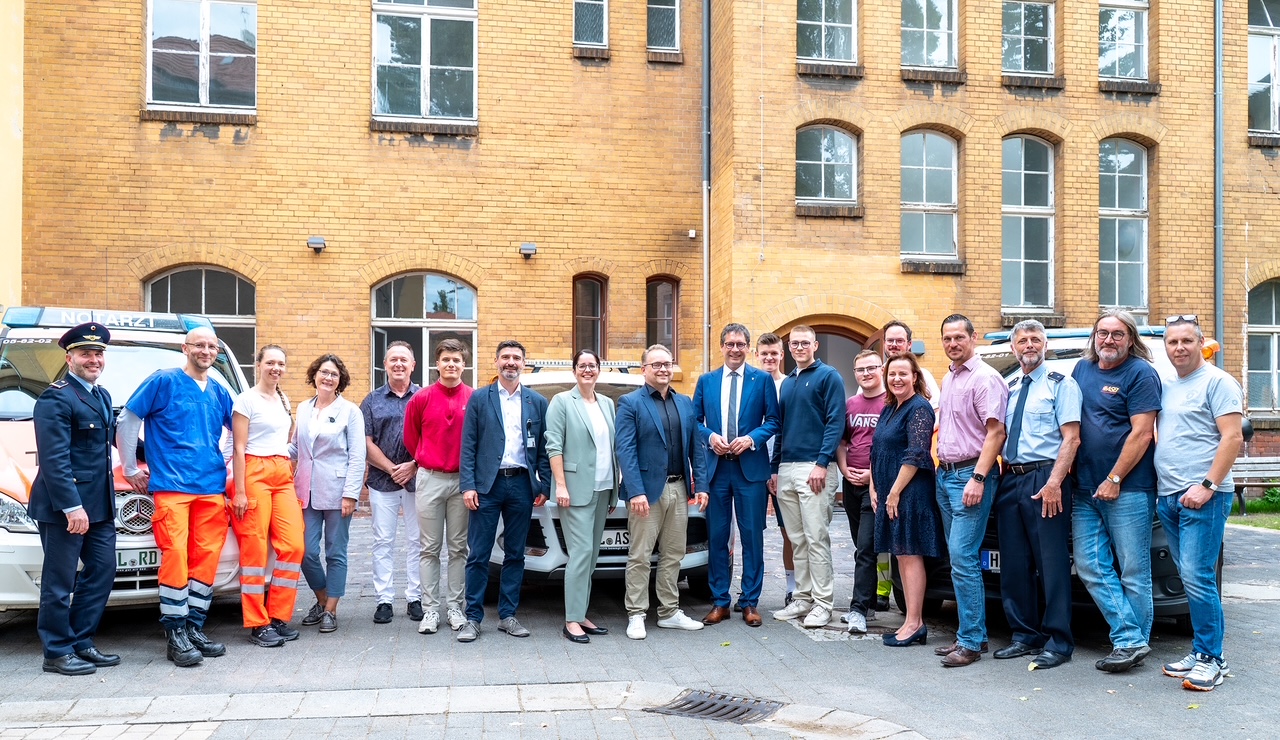 Gruppe von rund 20 Menschen steht vor  drei Rettungsfahrzeugen und einer Hausfassade.Hausfassade. Daru