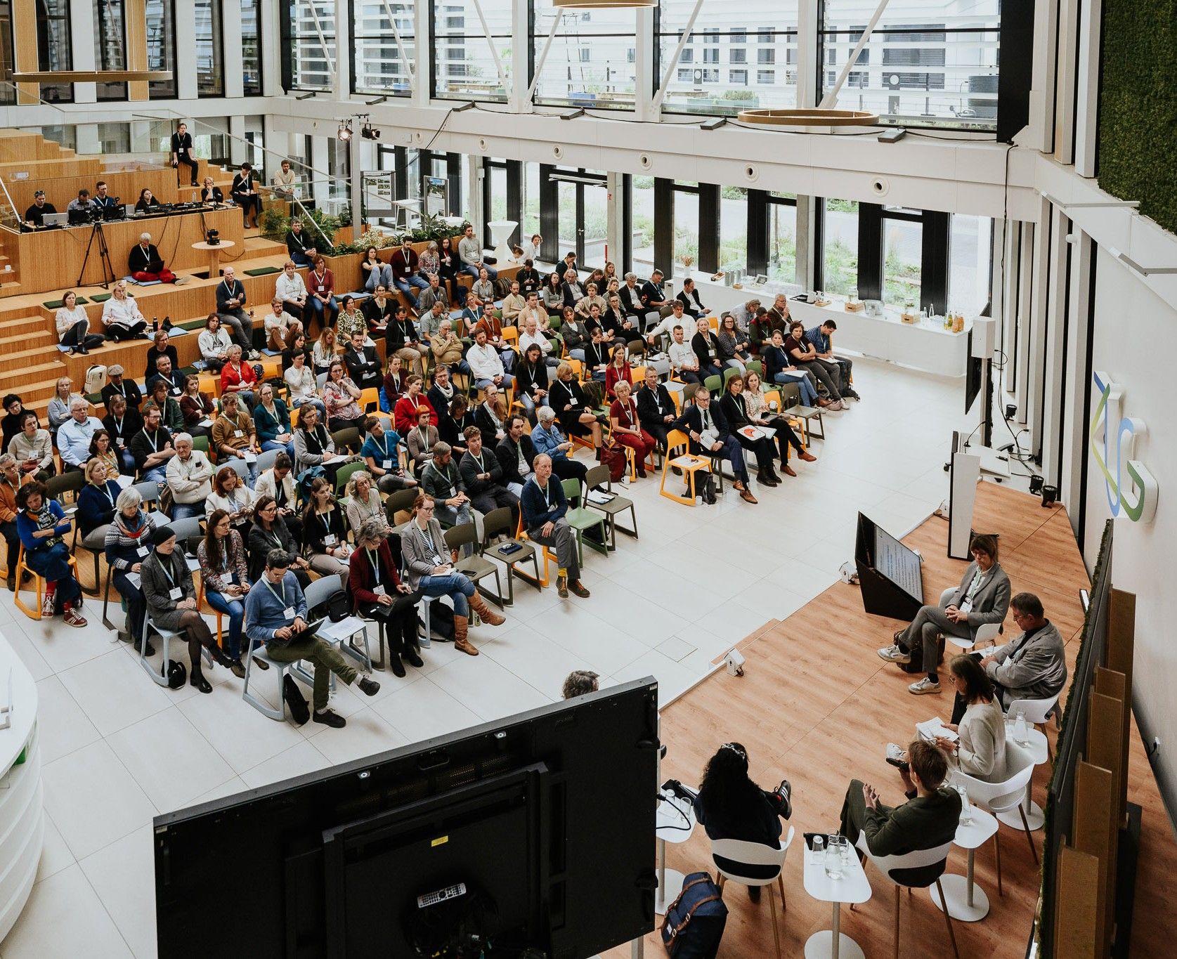 Menschengruppe sitzt in einem Hörsaal, Podiumsveranstalter auf einer Bühne moderieren zum Thema Natürlicher Klimaschutz