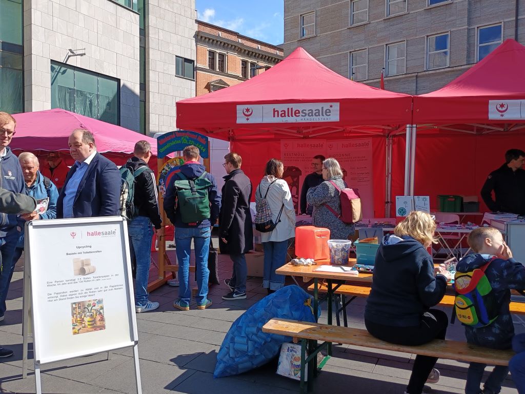 Auf einem Marktplatz stehen mehrere Pavillons. An der Sichtseite werden Aktionen angeboten. Ein Basteltisch steht im Fordergrund, ein Glücksrad weiter hinten. Menschen sind in Gesprächen vertieft.