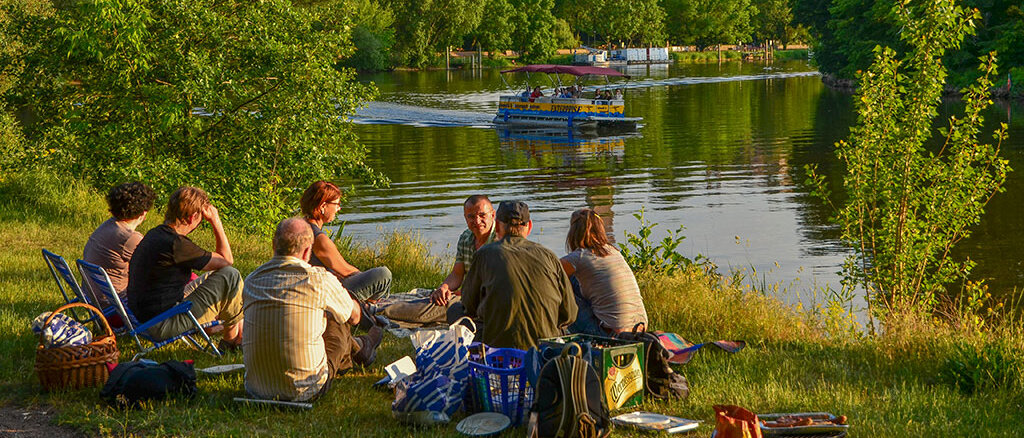 Eine Gruppe Personen sitzt am Saaleufer auf Decken zum Picknick, im Hintergrund die Burg Giebichenstein, 