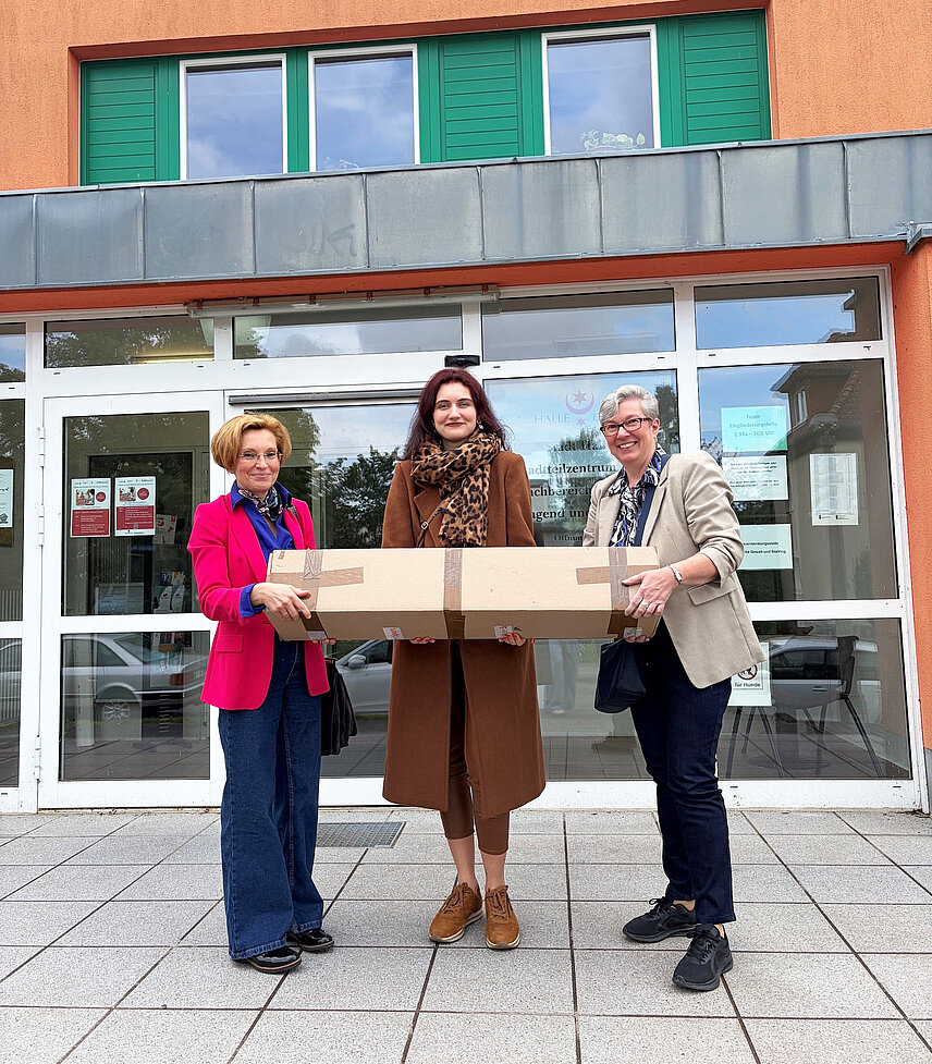 Drei Frauen halten gemeinsam ein Paket. Sie stehen vor dem Eingang eines Hauses.
