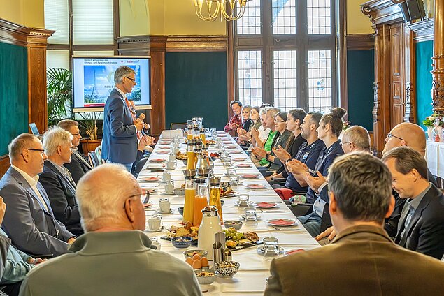 Eine lange Tafel mit Essen und Getränken. Zwischen den sitzenden Personen steht der Bürgermeister und spricht.