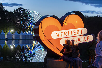 Zwei Personen sitzen vor dem Verliebt in Halle Logo, im Hintergrund das Riesenrad.