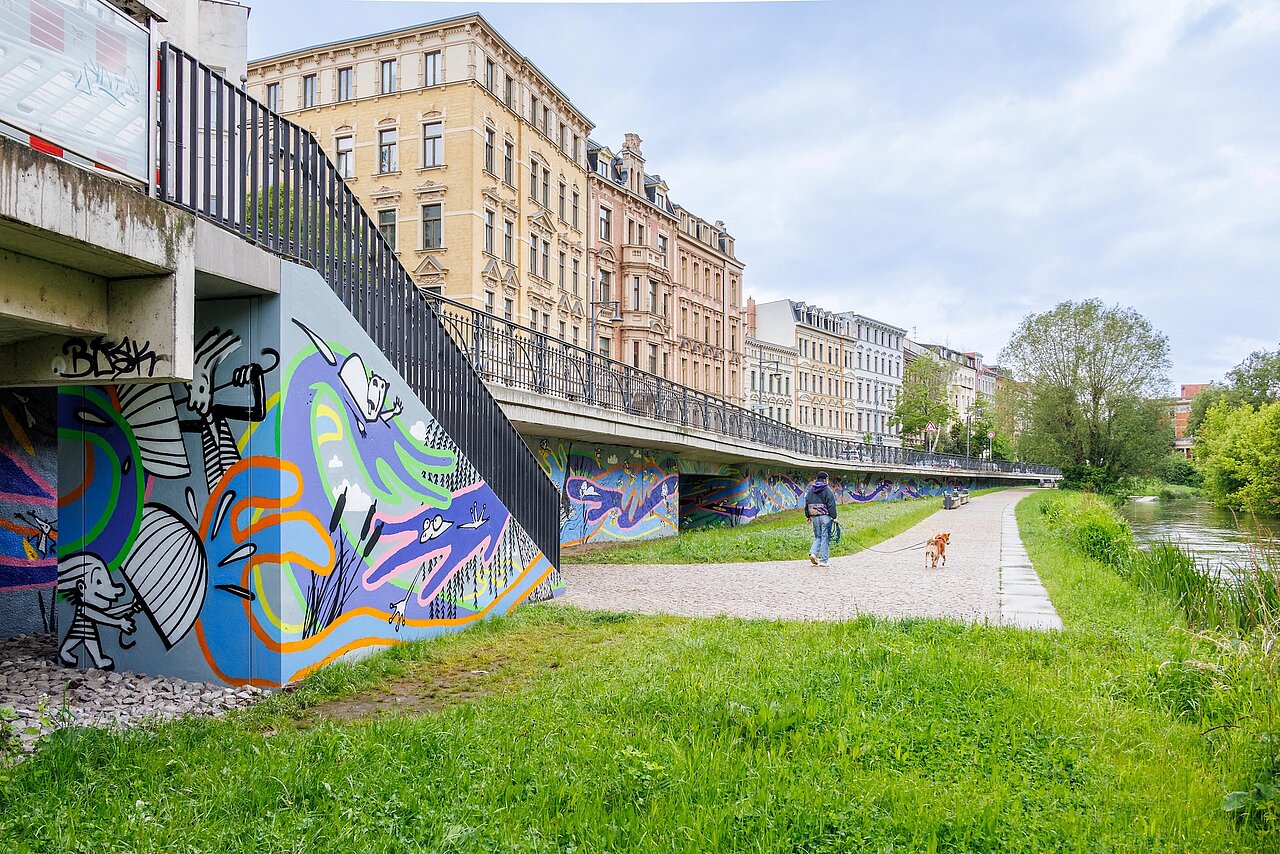 Blick auf eine bunt gestaltete lange Mauer (linsk), die das Areal des Saalearms Mühlgraben (rechts) begrenzt. 