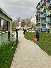 Im Hintergrund sieht man Erwachsene und Kinder mit Müllsäcken in der Hand die Müll auf einer Grünfläche auflesen.