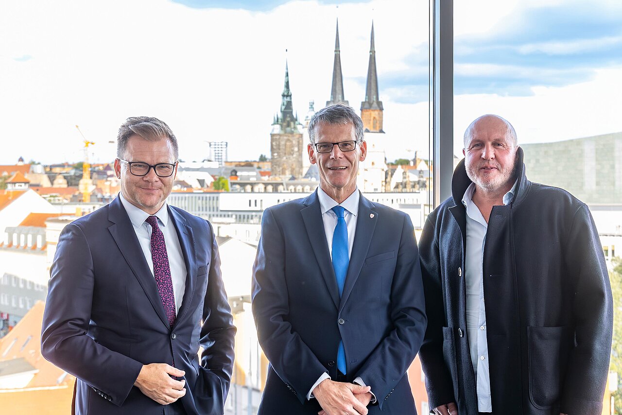 Drei Personen stehen vor einem Fenster, dahinter die Altstadt von Halle.