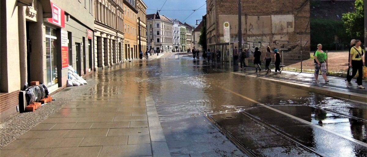 Hochwasser strömt im Gleisbett einer Straßenbahn, bedeckt Straße und Fußweg, rechts und links Häuser, Sandsäcke vor Eingängen 