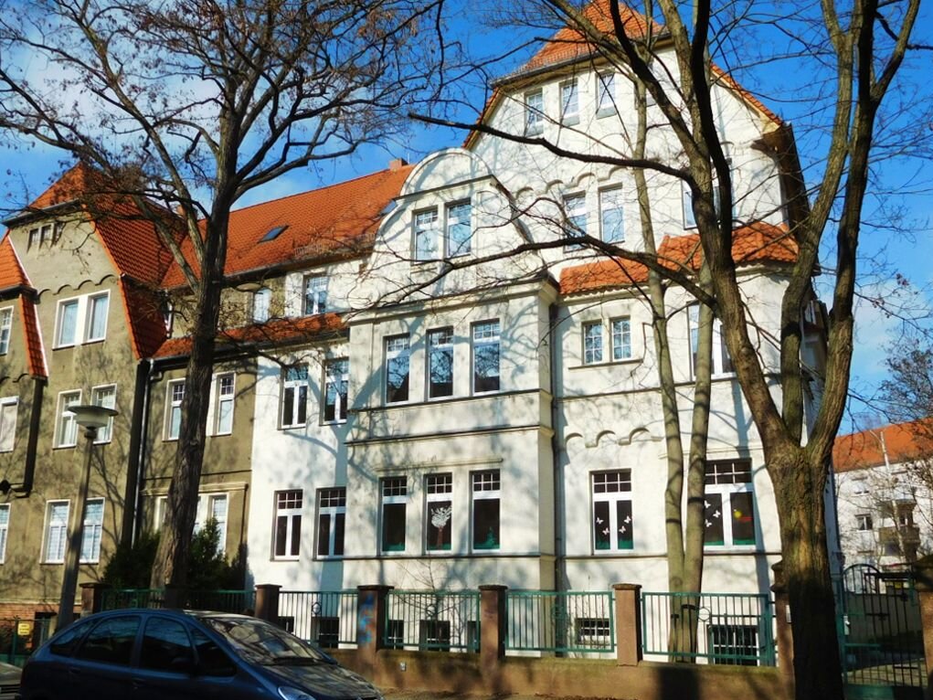 dreigeschossiges Gebäude mit vielen Fenstern und heller Fassade, zum Fußweg abgegrenzt durch Metallzaun mit Steinsockel, rechts und links davor auf dem Fußweg stehen  Bäume, links im vordergrund steht ein parkendes Auto auf der Straße