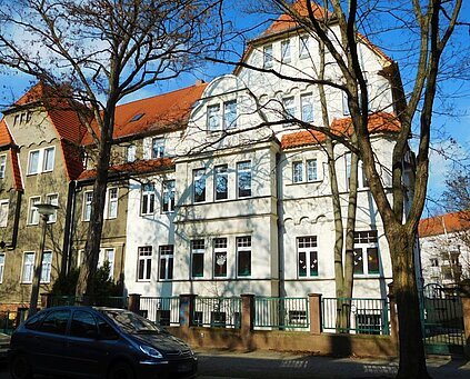 dreigeschossiges Gebäude mit vielen Fenstern und heller Fassade, zum Fußweg abgegrenzt durch Metallzaun mit Steinsockel, rechts und links davor auf dem Fußweg stehen  Bäume, links im vordergrund steht ein parkendes Auto auf der Straße