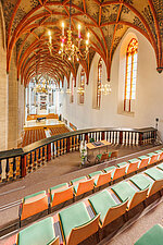 Blick von der Kirchenempore mit Bestuhlung in den untenliegenden Kirchenraum in Richtung des ehemaligen Altarbereichs, in dem ein Flügel aufgebaut und eine Orgel fest installiert ist. Im Vordergrund der Blick auf die reich verzierten Deckenbögen des Kirchenraumes und die strahlenden Deckenleuchter. Im rechten Bildrand die langgestreckten Kirchenfenster.rahlen. Am rechten oberen Bildrand die barocke Kirchenorgel