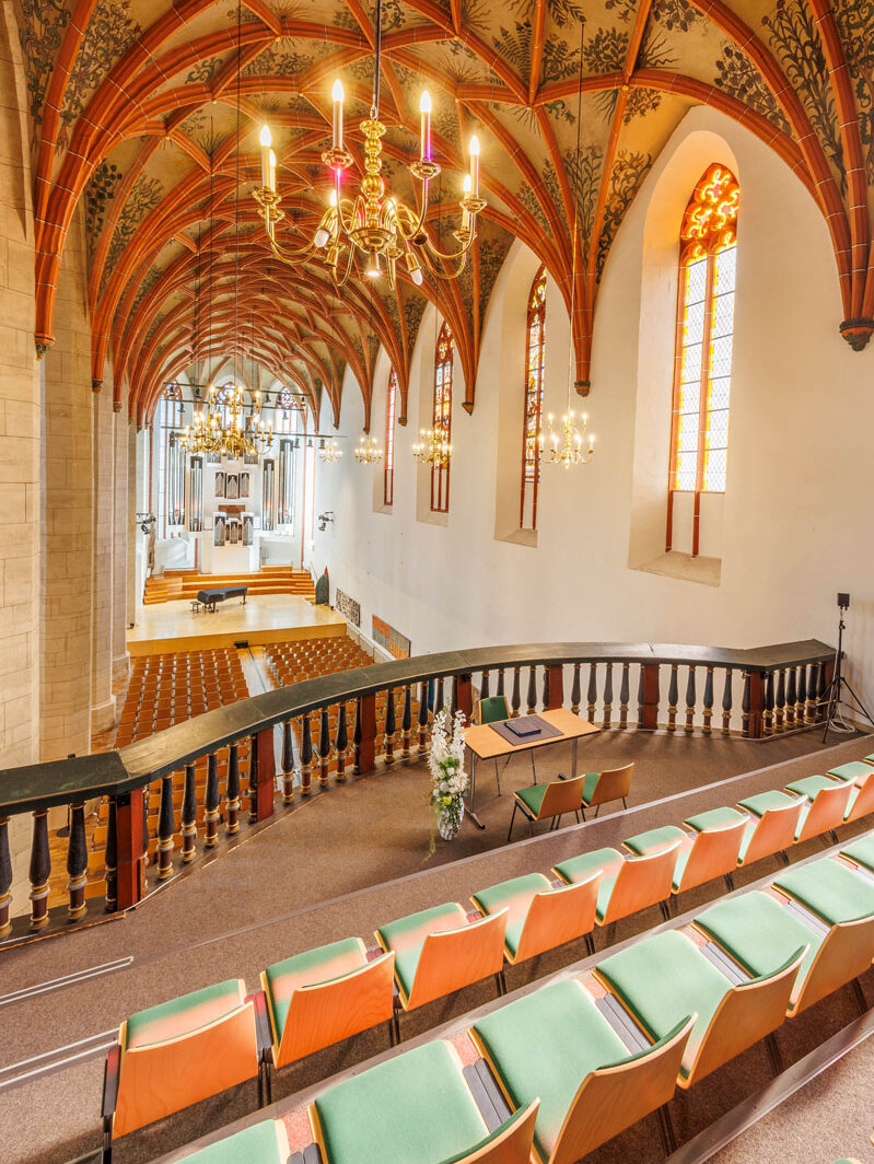 Blick von der Kirchenempore mit Bestuhlung in den untenliegenden Kirchenraum in Richtung des ehemaligen Altarbereichs, in dem ein Flügel aufgebaut und eine Orgel fest installiert ist. Im Vordergrund der Blick auf die reich verzierten Deckenbögen des Kirchenraumes und die strahlenden Deckenleuchter. Im rechten Bildrand die langgestreckten Kirchenfenster.rahlen. Am rechten oberen Bildrand die barocke Kirchenorgel