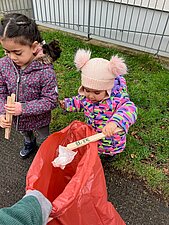 Zwei kleine Mädchen halten Holzzangen in der Hand. Ein Mädchen legt damit Müll in den Müllsack.