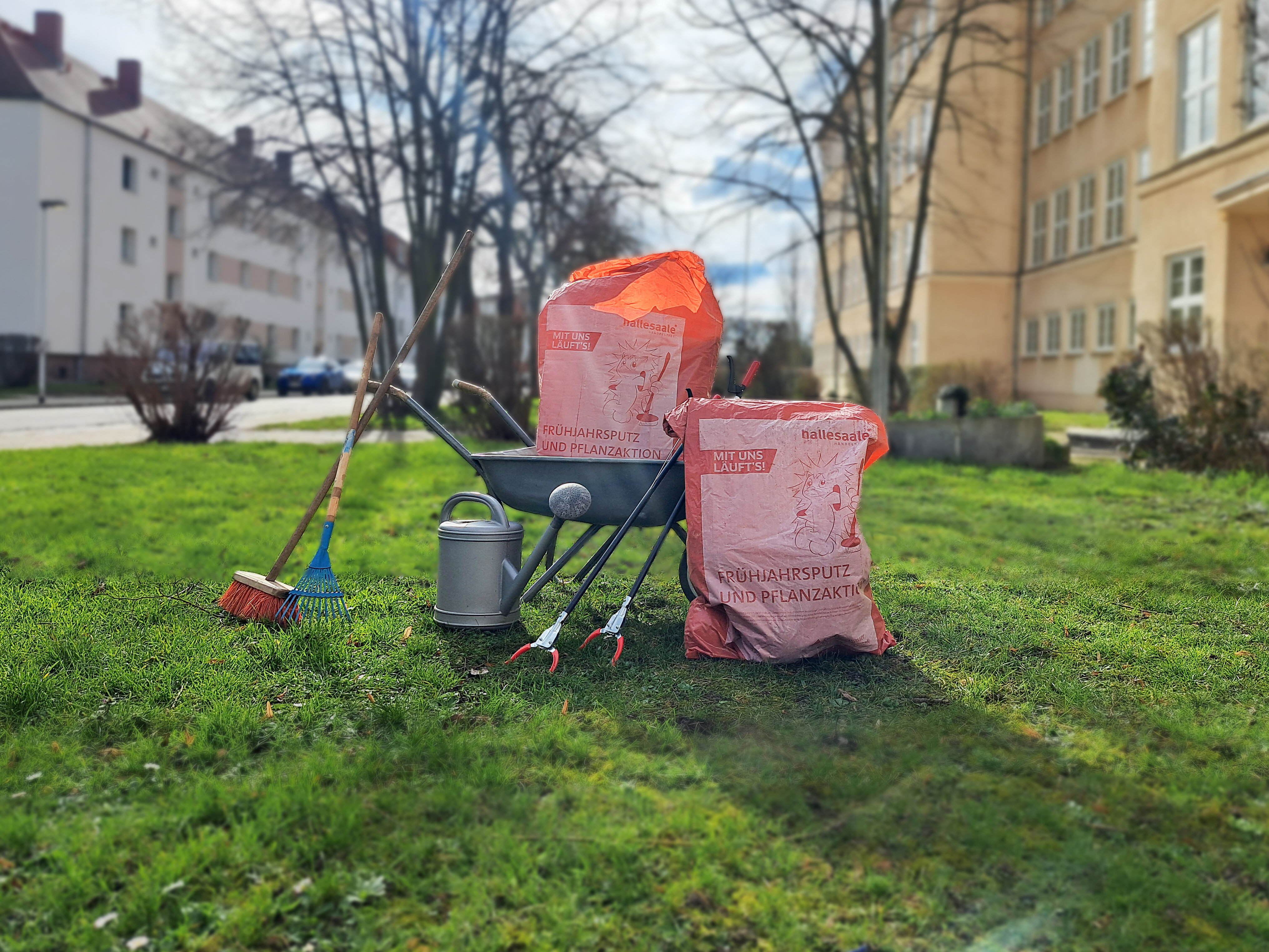 Frühjahrsputzsäcke platziert auf einer Wiese mit Schubkarre, Besen und Greifzangen