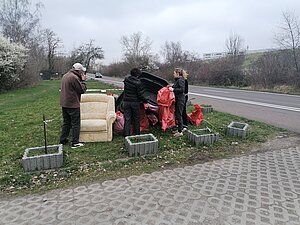 Foto zeigt vier Personen, die gesammelten Müll auf einer Grünfläche abstellen und Müll in einen Müllsack geben.