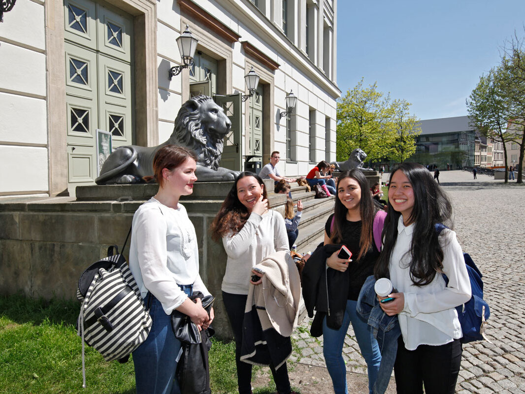 mehrere Studentinnen internationaler Herkunft stehen auf dem Uniplatz vor dem Löwengebäude
