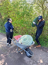 2 Personen bergen Müll aus den Büschen, im Vordergrund steht eine Schubkarre befüllt mit Müll.
