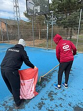 Eine Person kehrt den Dreck auf dem Spielfeld zusammen und die andere Person hält den Müllsack bereit.