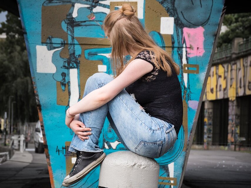 Ein jugendliches Mädchens hockt auf einem Poller mit abgewendetem Gesicht, im Hintergrund eine Graffiti-Wand