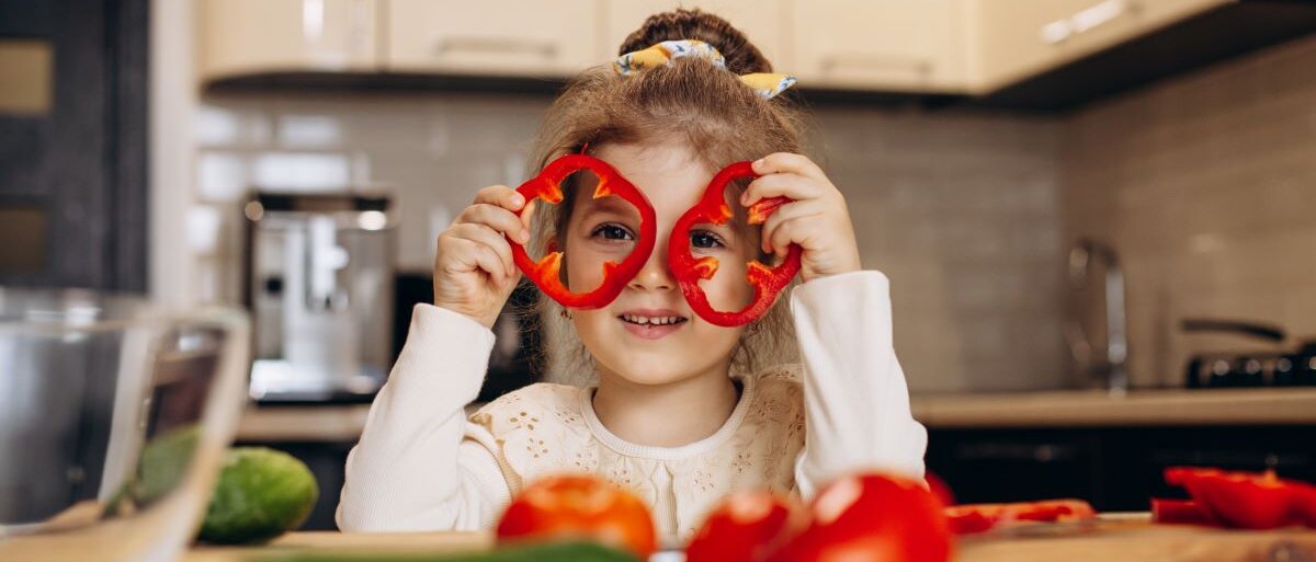 Ein kleines niedliches Mädchen bereitet Salat aus frischer Paprika zu und hält sich Paprikaringe als Brille um die Augen.. 