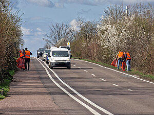 Mehrere Mitarbeitende laufen mit Warnwesten, Greifzangen und Müllsäcken am Straßenrand entlang und sammeln Streumüll auf.