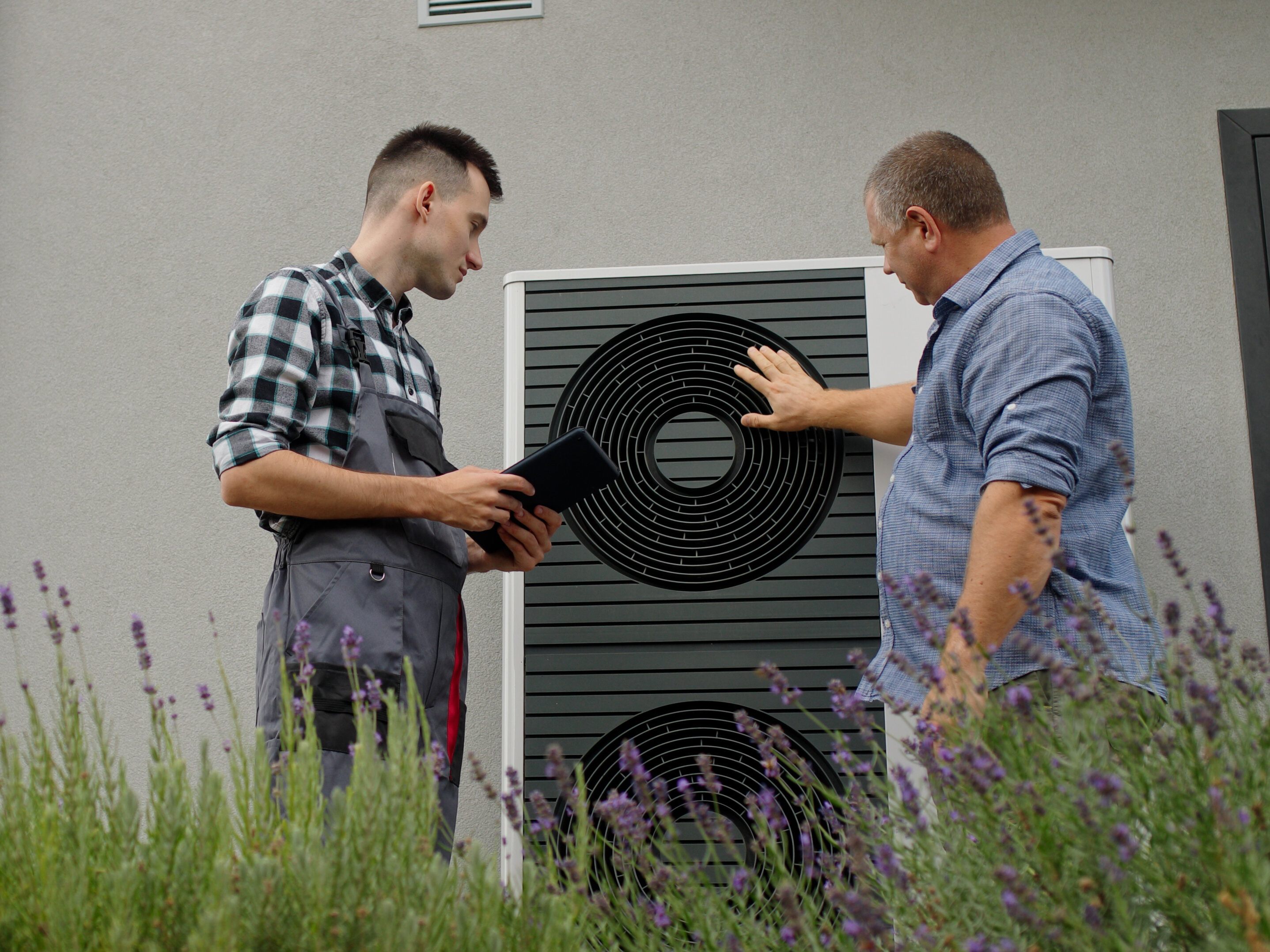 Zwei Männer (einer HAndwerker) betrachten eine Wärmepumpe vor einer Hausfassade