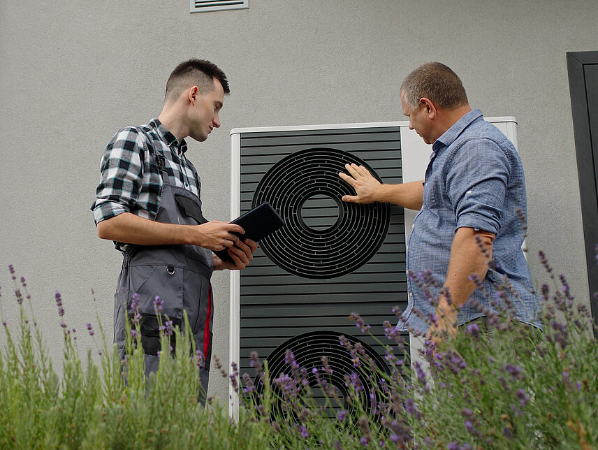 Zwei Männer (einer HAndwerker) betrachten eine Wärmepumpe vor einer Hausfassade