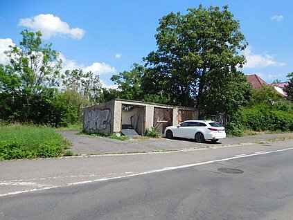 mehrere alte Garagen an einer Straße, im Hintergrund Bäume, davor parkt ein Auto