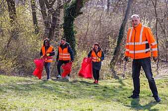 Ein Mitarbeiter ist rechts im Bild mit einer Greifzange in der Hand zu sehen und im Hintergrund befinden sich 3 Mitarbeitende mit Warnwesten, die Streumüll aufsammeln.