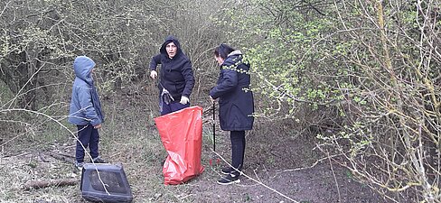Zwei Frauen und ein Kind sammeln Streumüll auf. Im Vordergrund ist ein roter Müllsack zu sehen.