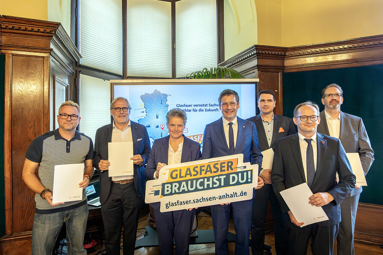 Oberbürgermeister Dr. Alexander Vogt und Digital-Ministerin Dr. Lydia Hüskens halten ein Schild mit der Aufschrift: „Glasfaser brauchst du!". links und rechts stehen fünf Männer mit unterzeichneten Verträgen in dden Händen