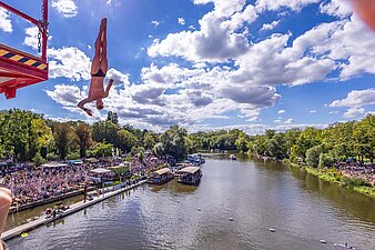 ein Springer in der Luft, an den Ufern der Saale sitzen viele Leute, um zuzuschauen