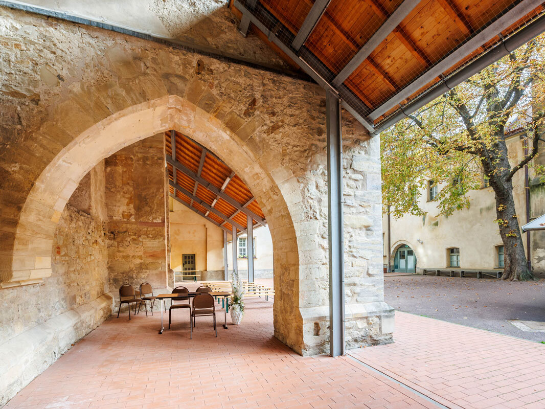 Bild eines überdachten sandsteinernen Torbogens in einem Innenhof mit roten Pflastersteinen, im Hintergrund Tische, Stühle und Bänke für eine Trauung. Sowie ein Kastanienbaum an der Seite des Bildes.