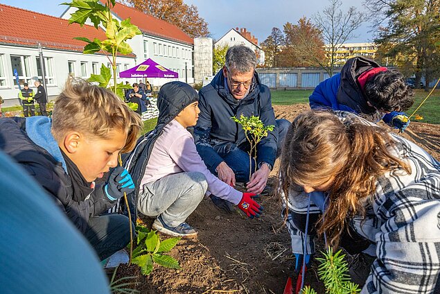 Der Bürgermeister kniet neben einem neu eingepflanzten Baum vor derm Schulgebäude.