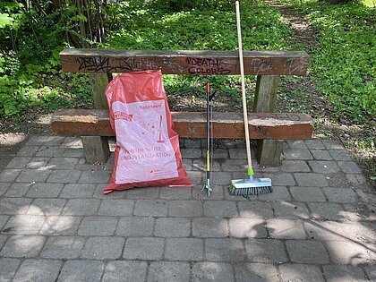 Foto von befülltem Müllsack vor einer Holzbank platziert und Müllzangen daneben