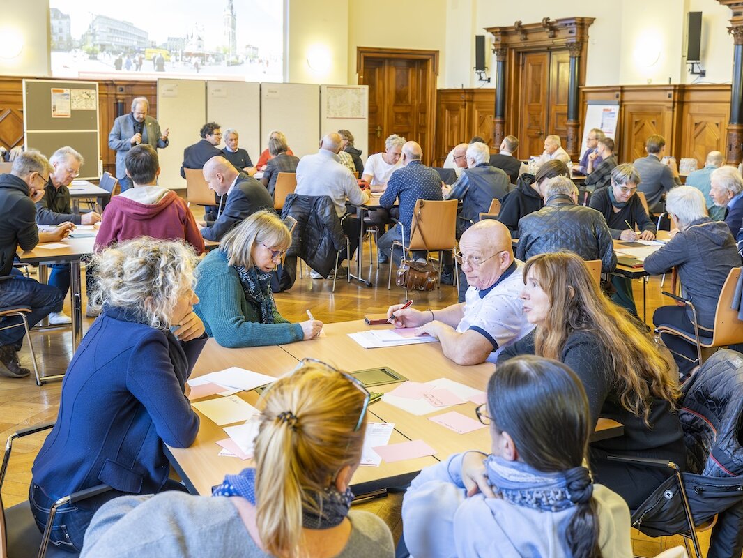 Blick in den großen Saal im Stadthaus. Bürgerinnen und Bürger sitzen in Gruppen an mehreren Tischen und überlegen sich Antworten auf 3 FragenGruppen an Tischen 