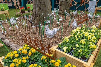 weiße Hühnerfiguren mit gebastelten Blumen an einem Baum, im Vordergrund Blumenkästen mit gelben Blumen