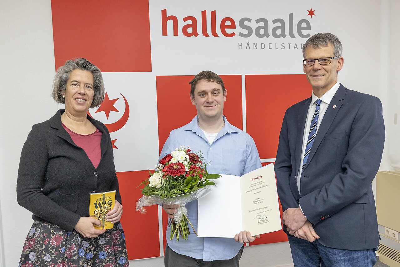 Zu sehen sind Innenministerin Dr. Tamara Zieschang, Hannes D., Bürgermeister Egbert Geier mit einem Strauß Blumen und einer Ukrunde vor dem Logo der Stadt Halle 
