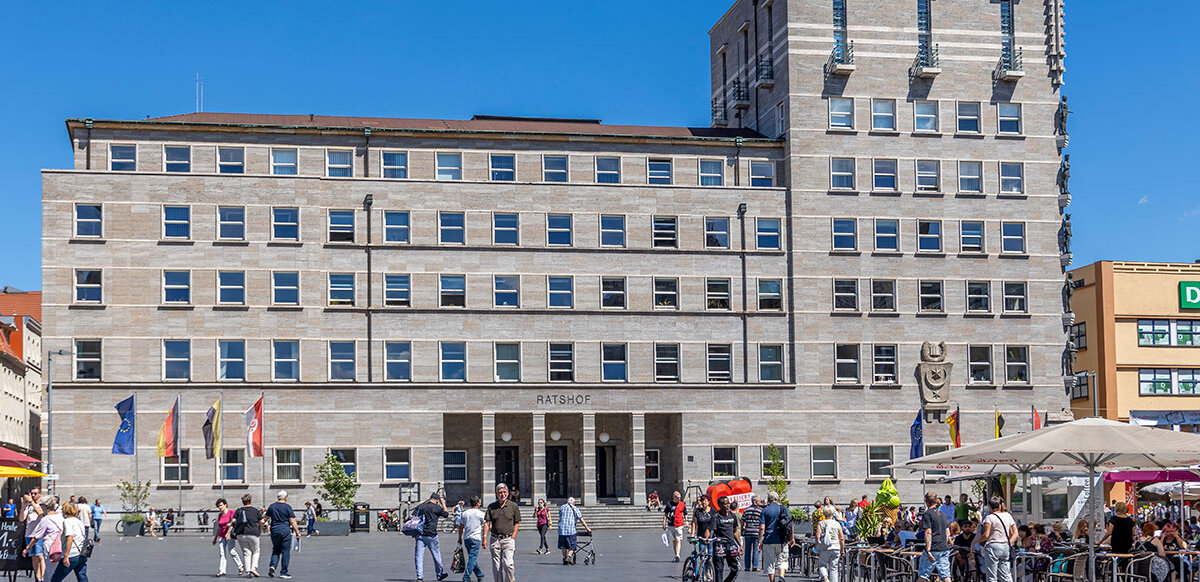 Blick auf das Ratshofsgebäude vom Marktplatz aus, davor laufen Menschen oder stehen an Marktständen
