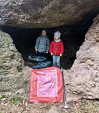 2 Kinder, die zwischen Felsen stehen. Im Vordergrund liegt ein roter Müllsack.