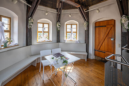 Weißer kleiner Raum im inneren eines Turmes, an den Wänden weiße Sitzbänke, in der Mitte ein kleiner weißer Tisch mit zwei Stühlen. Im Hintergrund drei Kreuzsprossenfenster aus hellem Holz sowie einer massiven hellen Holztür. Die Fensterbänke sind festlich geschmückt