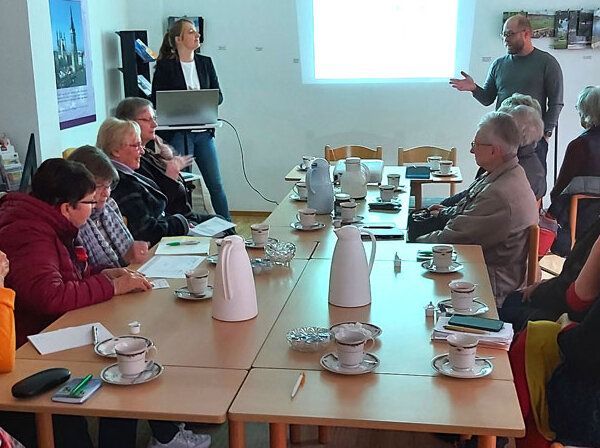 12 ältere Menschen bei einer Beratung an einem Tisch diskutieren anhand einer Beamerpräsentation