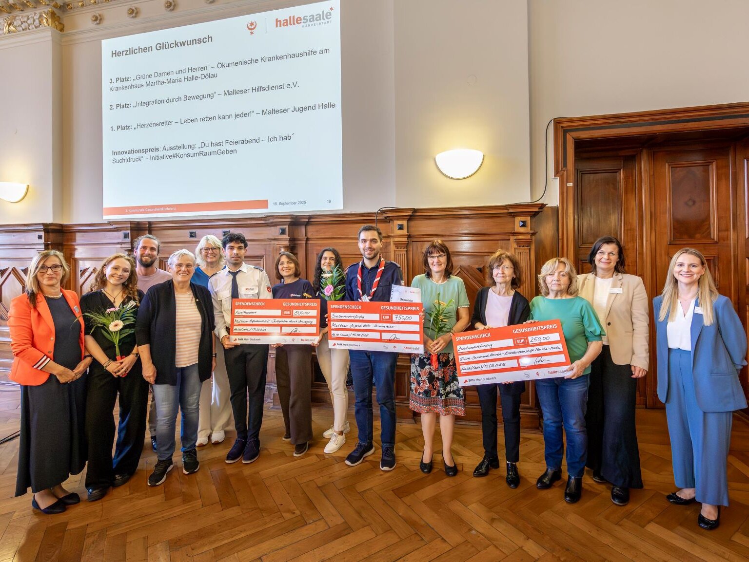 Preisverleihung 2025: Die drei besten Projekte mit Schecks, gemeinsam mit der Beigeordneten Frau Brederlow, der Leiterin des Fachbereichs Gesundheit Frau Dr. Gröger sowie der Koordinatorin für Gesundheitsförderung und Prävention Frau Anne Cierpinski.—Hintergrundfolie „Herzlichen Glückwunsch – Top 3