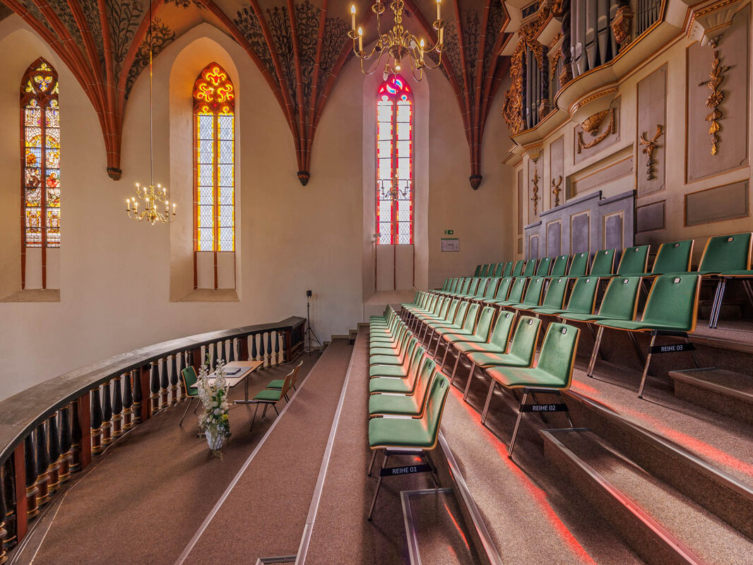 Bild einer Kirchenempore mit Bestuhlung auf vier abgestuften Ebenen, im Hintergrund die verzierten Deckenbögen der Kirche und drei Kirchenfenster, die im Licht der einfallenden Sonne bunt strahlen. Am rechten oberen Bildrand die barocke Kirchenorgel