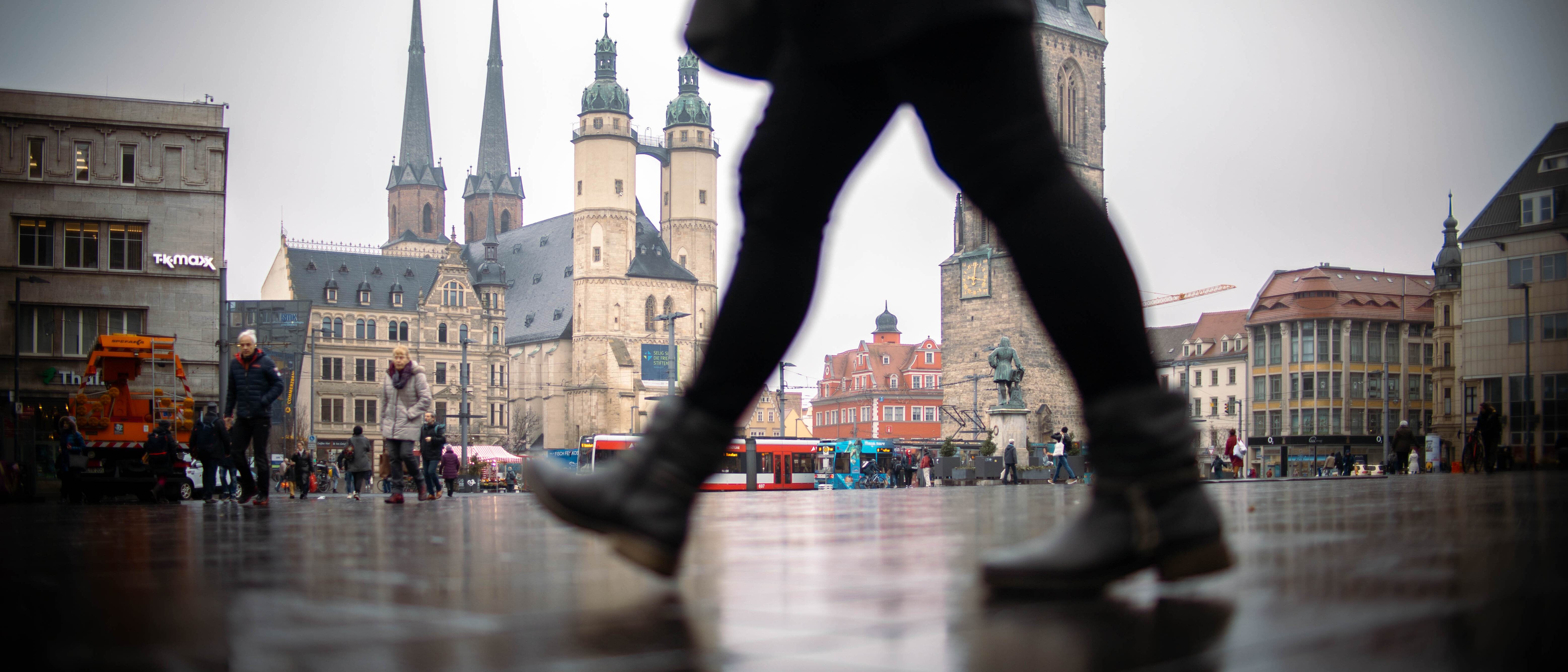 Bild in Froschperspektive mit Blick auf den Marktplatz Halle (Saale) mit Marktkirche, Marktschlösschen, Roter Turm und Händel-Denkmal. Im Vordergrund läuft eine Person durchs Bild, im Hintergrund sind mehrere Personen sowie eine Straßenbahn zu sehen. 